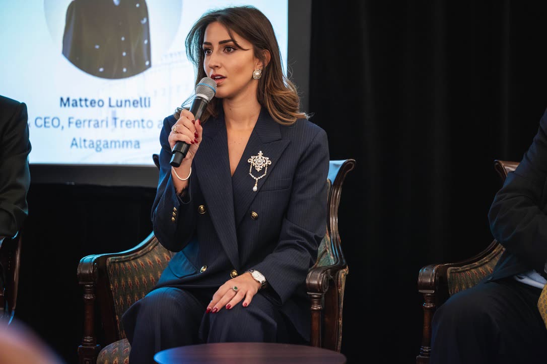 Camilla Brossa speaks into the microphone during a panel at the Italian Chamber of Commerce in London, in a navy pinstripe suit with gold buttons and a jewelled brooch