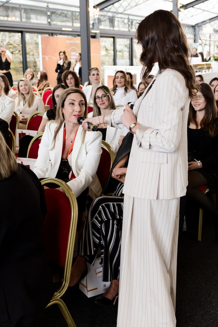 Camilla Brossa, seen from behind in a cream pinstripe suit, holds out the microphone to a woman seated in the audience