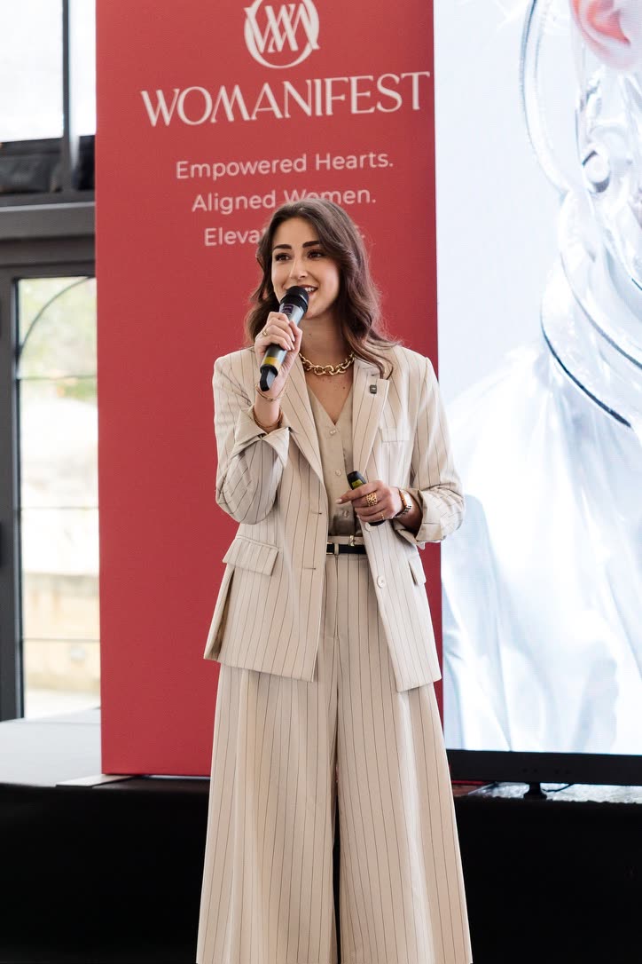Camilla Brossa full-length on the Womanifest stage, smiling into the microphone in a cream pinstripe suit and a gold chain necklace