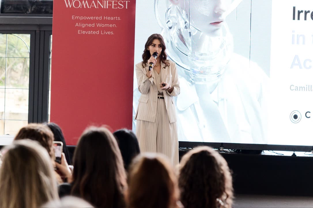 Camilla Brossa addresses the Womanifest audience in front of the screen showing the slide of her talk, in a cream pinstripe suit