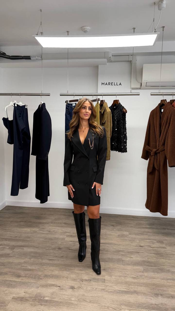 Camilla Brossa full-length in the Marella showroom, in a black double-breasted blazer dress with a gold chain and knee-high boots, in front of the rails