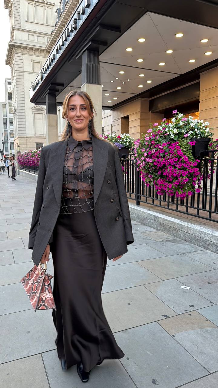 Camilla Brossa walking in London past a hotel entrance hung with flowers, in an oversized grey blazer, crystal top and long satin skirt, with a python-print bag