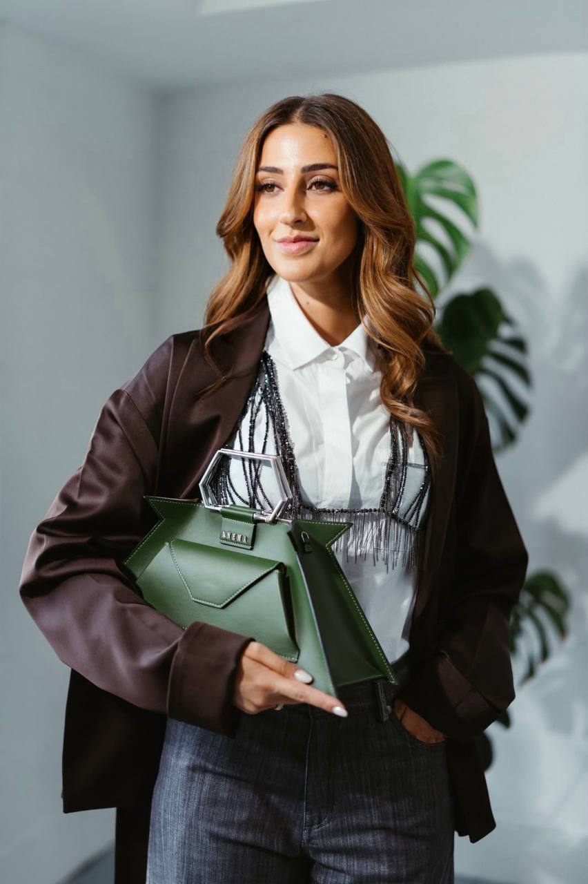 Camilla Brossa in a chocolate satin jacket over a white shirt, holding a green bag with a hexagonal handle