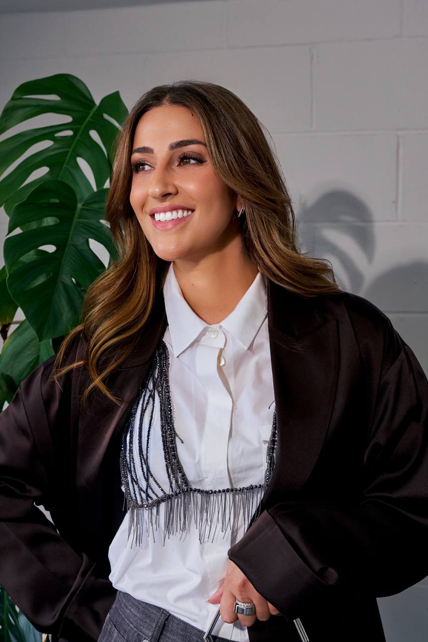 Close-up of Camilla Brossa smiling, in a satin jacket and crystal-fringe bralette, beside a monstera